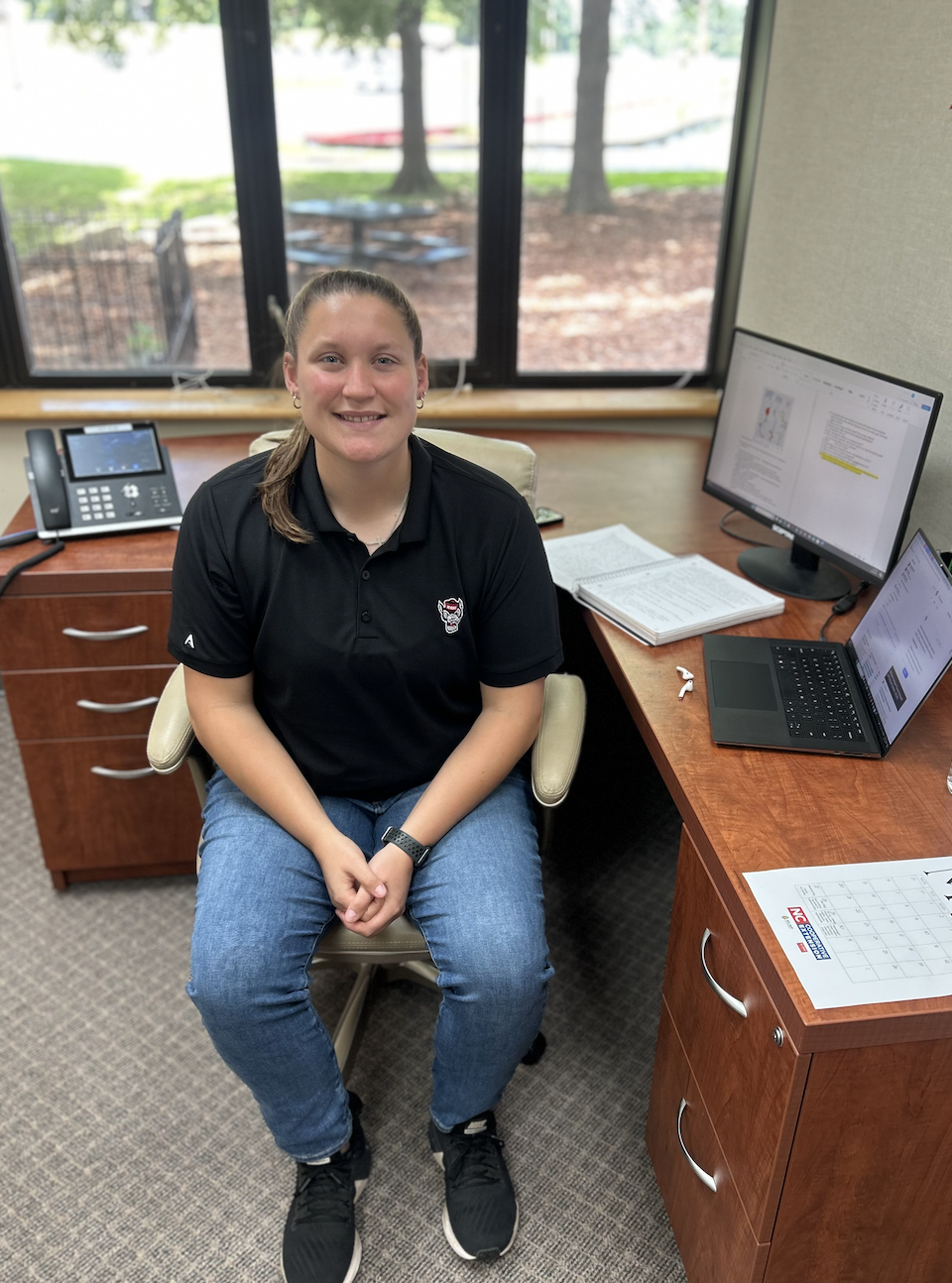 A woman in an NC State shirt sits at a desk.